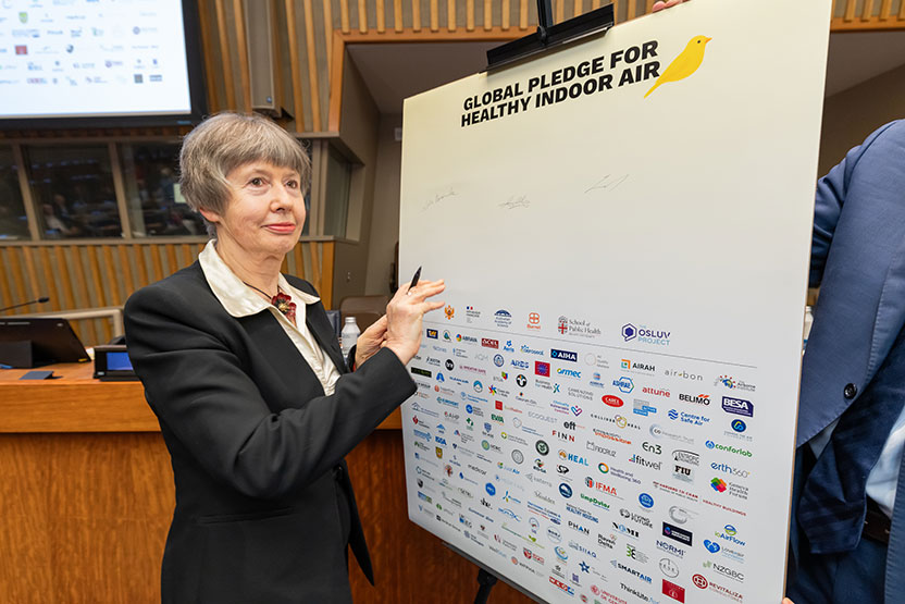 A woman in a black suit holds a pen up to a large posterboard that reads 'Global pledge for healthy indoor air' and features a yellow canary and dozens of colourful logos.